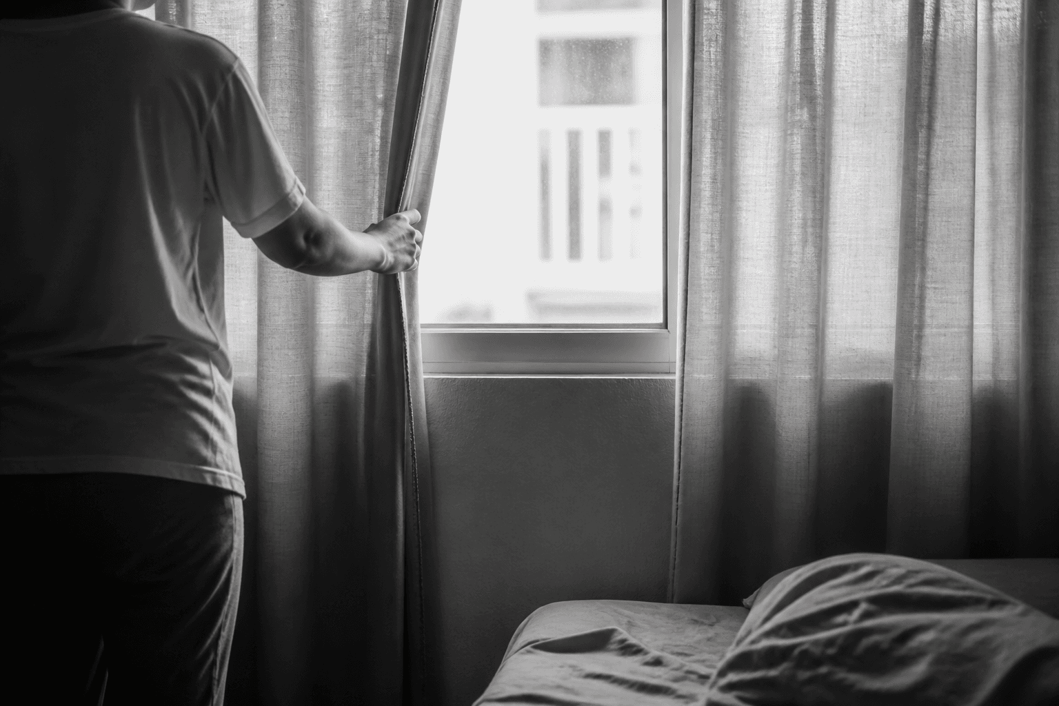 A person opens the curtains to morning light, with a muted-red fabric edge held against an otherwise black-and-white scene.