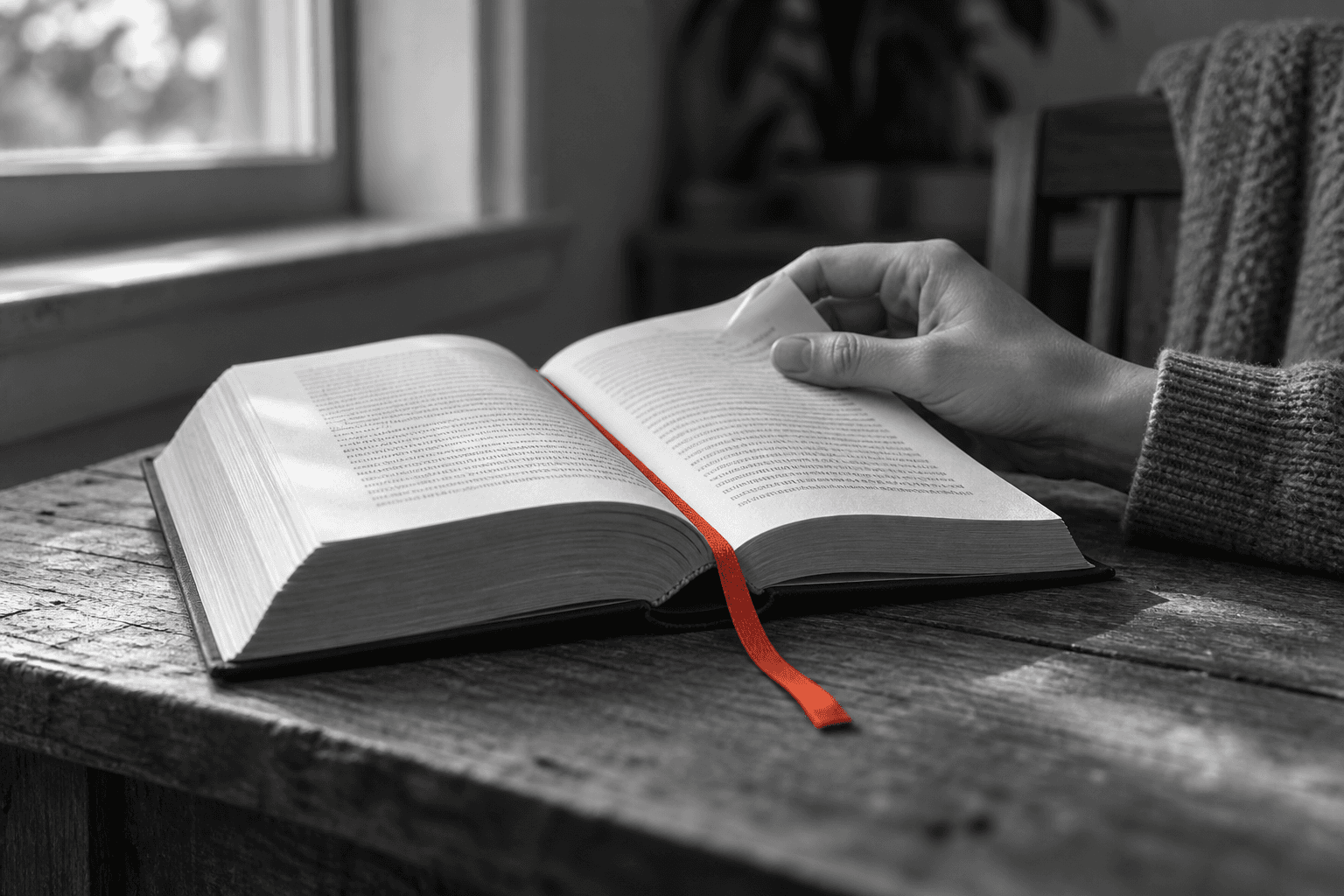 A hand opens a book at a red ribbon bookmark on a table beside a window, in an otherwise black-and-white photograph.